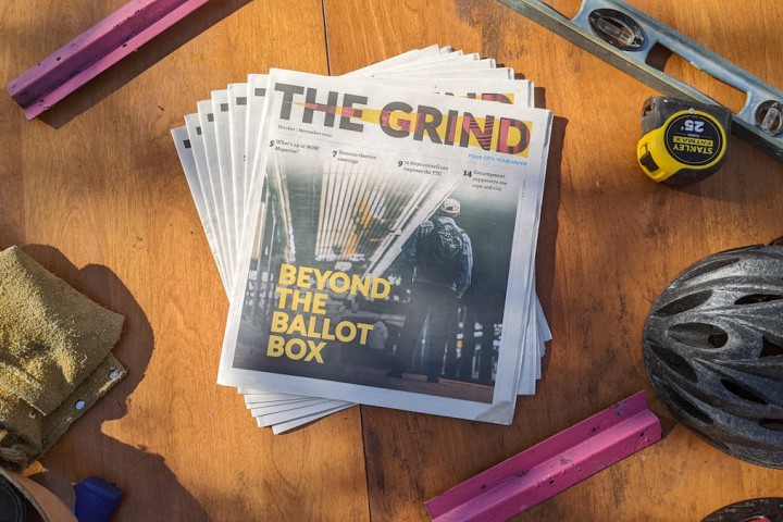 A photo taken from bird's-eye view. In the center, copies of an alt-weekly newspaper called The Grind are fanned out. The cover of The Grind shows a person standing in a subway station and the words