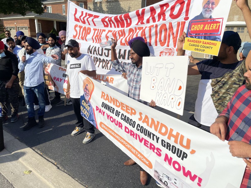 A photo of a crowd of brown men – some wearing turbans, some wearing baseball caps – holding a banner that says