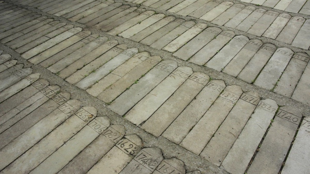 A photograph of gravestones on the grounds of Huronia. The gavestones are long, thin gray stones with only a number at the top. They are set into a concrete pad close together, nestled next to each other on the ground.