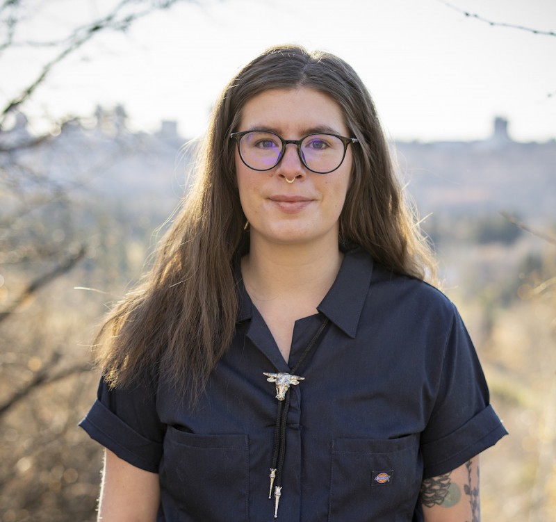 Jessica Johns, a light-skinned Cree woman, stands in front of a blurred fall backdrop and is looking straight at the camera. She has shoulder-length, wavy, light brown hair and blue eyes. She is wearing a navy blue collared jumpsuit accessorized with a black leather bolo tie with silver embellishments. She has a silver, triangular septum piercing and colourful tattoos of plants on her left arm.