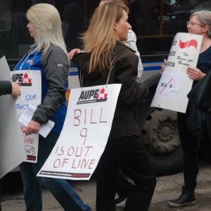 A person with long hair walks a picket line among others, wearing a sign that says